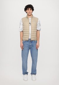 Beige puffer vest med glidelås foran, ribbet krage, brukt over en hvit, marmorert t-skjorte. Sammen med lyseblå jeans og hvite joggesko.