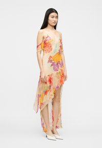 Jeune femme aux longs cheveux bruns portant une robe fleurie asymétrique et transparente avec des manches à épaules dénudées et des escarpins blancs à bouts pointus.