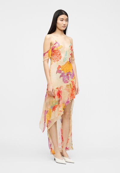 Νεαρή γυναίκα με μακριά σκουρόχρωμα μαλλιά, ντυμένη με διάφανο, ασύμμετρο φλοράλ φόρεμα με μανίκια χωρίς ώμους και λευκά μυτερά τακούνια.