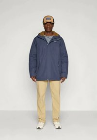 Σκούρο μπλε μπουφάν με επένδυση και κουκούλα με γούνα, μπροστινός φερμουάρ και κουμπιά. Φοριέται με ανοιχτό μπεζ παντελόνια και αθλητικά παπούτσια.