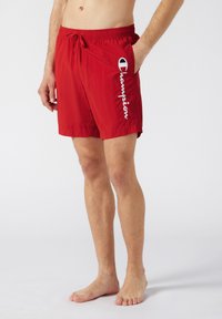 Un short de bain rouge en tissu léger. Il dispose d'une taille élastique avec cordon de serrage, d'une poche latérale et d'un détail logo "Champion" blanc.