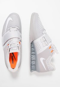 Grå træningssko med hvide detaljer, mesh-design og justerbar rem; har orange inderside og gennemsigtig hæl.