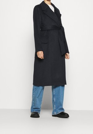 Personne portant un long manteau noir ceinturé sur un haut blanc, un jean large bleu et des chaussures noires, se tenant devant un fond clair.