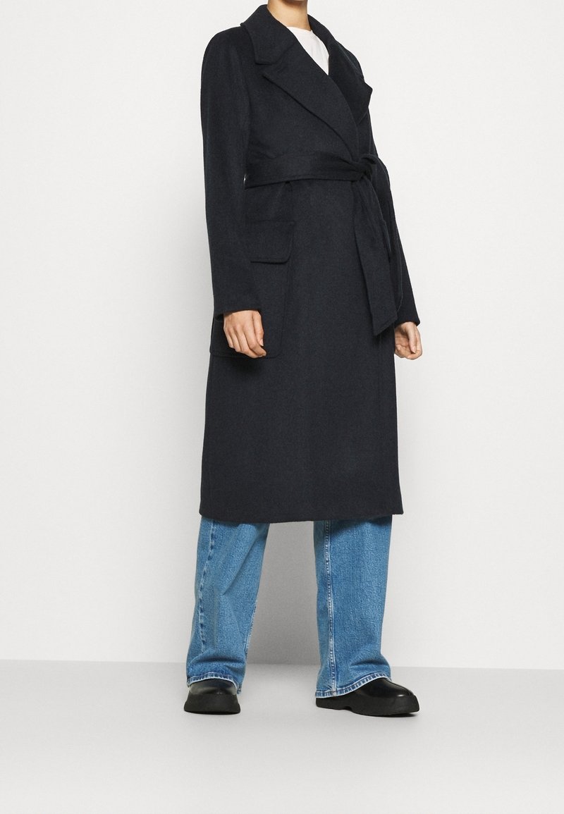 Persoon in een lange zwarte geknoopte jas over een witte top, blauwe wijde jeans en zwarte schoenen, staand tegen een effen lichte achtergrond.