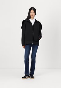 Veste noire zippée avec un design de col unique, poches avant, assortie à un jeans en denim et des baskets noires avec des accents beige.