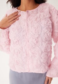 Rosa blommig jacka gjord av lager av tyg som liknar rosenblad. Har rund hals, långa ärmar och dragkedja fram.