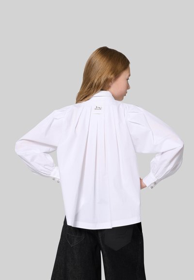White, long-sleeve shirt with a pleated back, buttoned cuffs, and a logo tag at the neckline. Worn with dark, wide-legged pants.