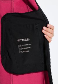 Intérieur d'une veste en polaire montrant une poche noire avec une fermeture éclair, affichant le logo "HYRAR" et des étiquettes indiquant résistant à l'eau, résistant au vent, respirant.