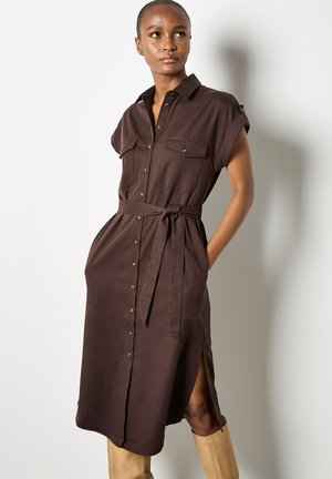 Robe chemise marron à manches courtes, avec deux poches poitrine, une taille ceinturée, un bouton sur le devant et des fentes latérales. Tissu lisse avec une finition mate.