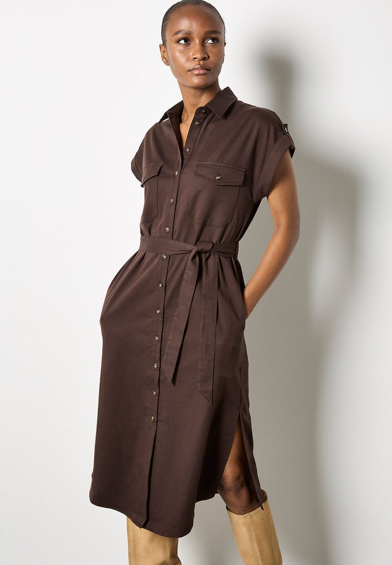 Robe chemise marron à manches courtes, avec deux poches poitrine, une taille ceinturée, un bouton sur le devant et des fentes latérales. Tissu lisse avec une finition mate.
