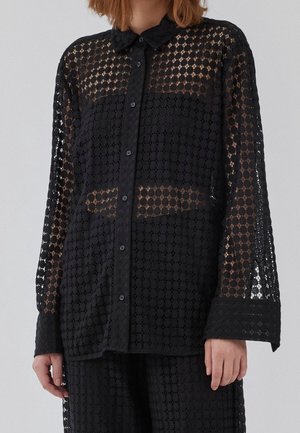 Person wearing a black sheer long-sleeve button-up shirt with a geometric lace pattern and matching black pants.