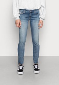 Persona che indossa jeans skinny azzurri chiari con fessure alla caviglia e sneakers bianche e nere, in piedi contro un muro e un pavimento neutri.