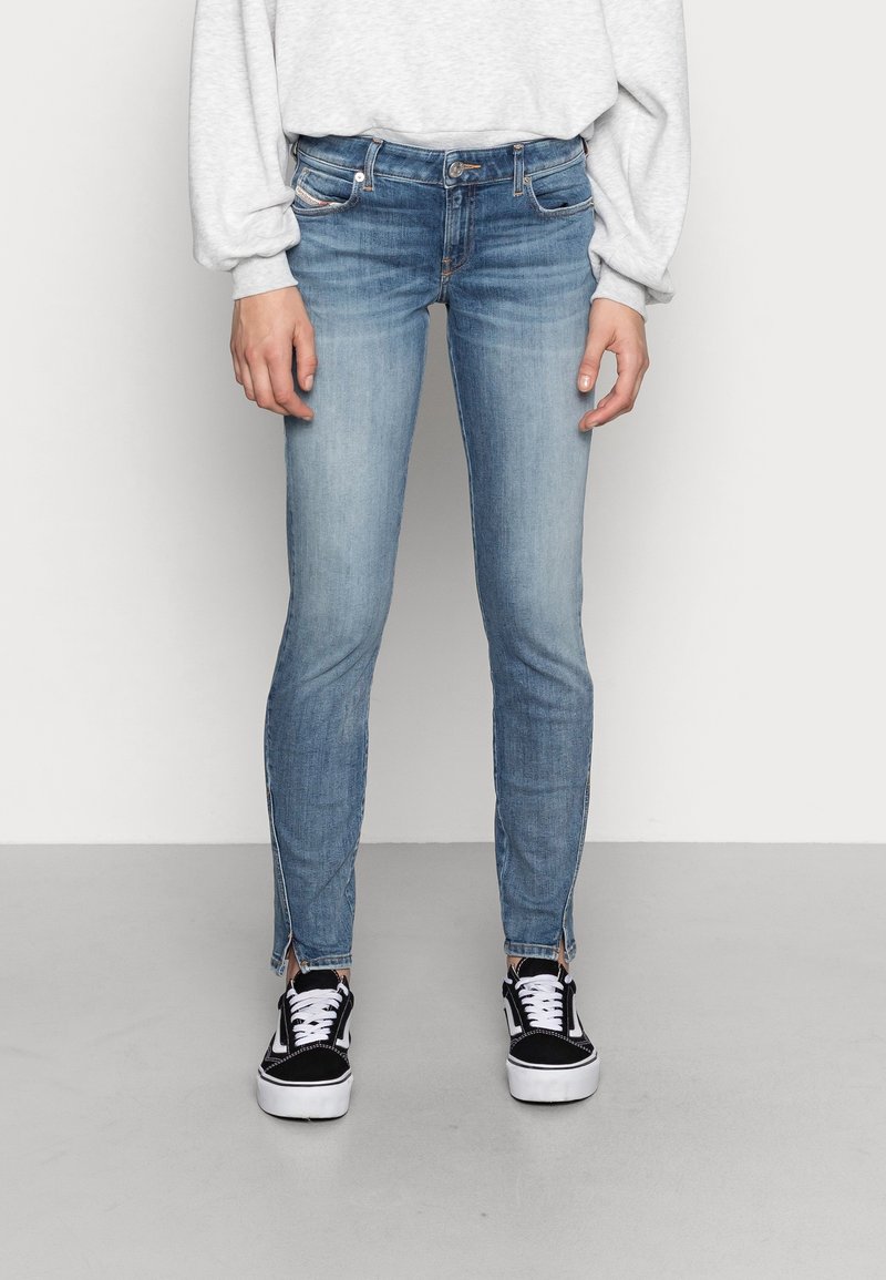 Persona che indossa jeans skinny azzurri chiari con fessure alla caviglia e sneakers bianche e nere, in piedi contro un muro e un pavimento neutri.