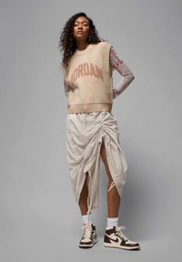 Beige strikket ermeløs genser med "JORDAN" i brune bokstaver, kombinert med et kremfarget rynket skjørt og brune høyde sneakers.