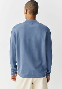 Jasnoniebieski sweter z długim rękawem i kołnierzykiem, o gładkiej fakturze i luźnym kroju, wykończony subtelnym detalem wzdłuż hemline'u.