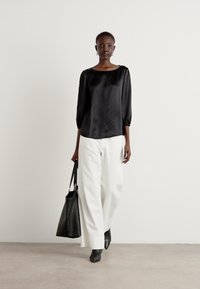 Zwarte satijnen blouse met driekwartmouwen, gecombineerd met witte wijde broek. Model draagt een zwarte tote tas en heeft enkelboots aan.