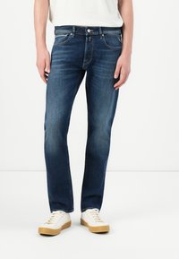 Person wearing dark blue straight-fit jeans and white sneakers with gum soles, standing against a plain white background.