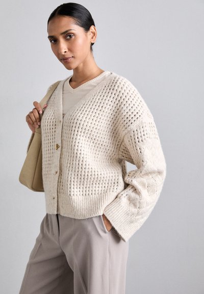 Cardigan texturat de crem cu guler în V, închidere cu nasturi și margini cu rădăcină, asortat cu un top ușor și pantaloni taupe.