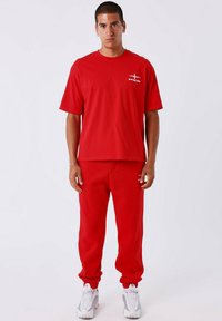 T-shirt rossa in cotone con maniche corte e un piccolo logo sul petto, abbinata a pantaloni della tuta rossi con polsini elasticizzati. Le sneakers bianche completano l'outfit.