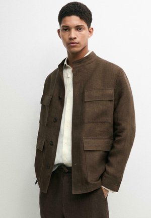 Giovane uomo con giacca marrone con tasche a patta sopra una camicia bianca con bottoni, in piedi davanti a uno sfondo chiaro e uniforme.