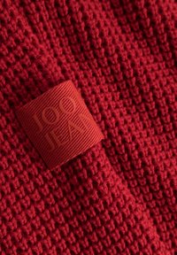 Zbliżenie na czerwoną dzianinę z czerwoną wplecioną metką z nazwą marki "JOOP! JEANS."
