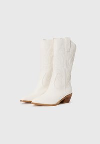 Bottes de cowboy blanches avec des motifs en relief en forme de tourbillon, bouts pointus et un petit talon empilé. Fabriquées en matériau texturé avec des coutures durables.