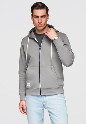 Jonge man in een grijze hoodie met rits en witte trekkoorden en lichtblauwe spijkerbroek, die zijn kraag rechtzet, neutrale grijze achtergrond.
