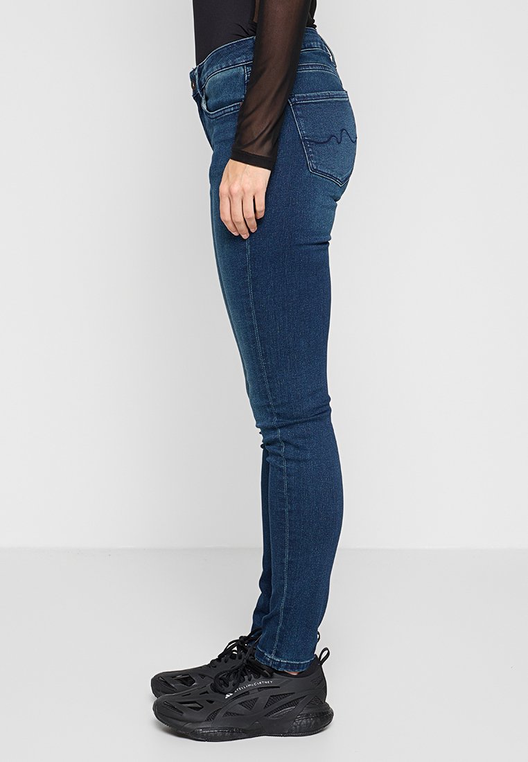 Jeans skinny blu con lavaggio medio, caratterizzati da tasche posteriori con cuciture curve, indossati con sneaker sportive nere.