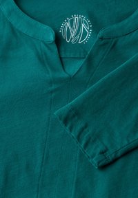 Türkises Shirt mit V-Ausschnitt und langen Ärmeln, gefertigt aus weichem Stoff. Verfügt über dezente Nähte und ein kreisförmiges Logo am Kragen.