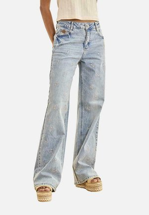 Jeans azul claro de pierna ancha con patrones bordados de estrellas, combinados con sandalias beige de plataforma y un top corto sin mangas en color crema.