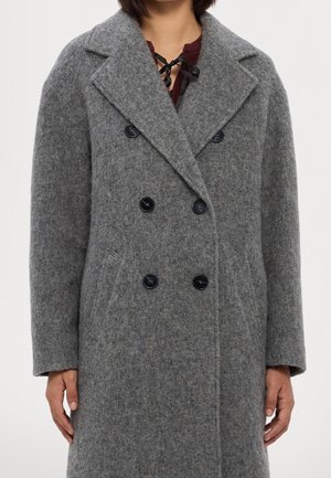 Osoba ubrana w długi szary dwurzędowy wełniany płaszcz z szerokimi klapami i czarnymi guzikami, ręce swobodnie opuszczone wzdłuż ciała.