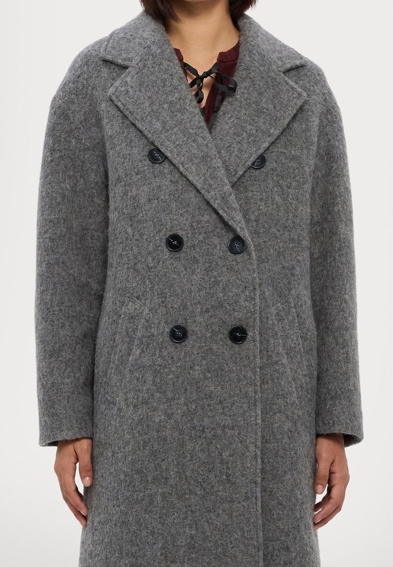 Personne portant un long manteau en laine gris à double boutonnage avec de larges revers et des boutons noirs, mains détendues sur les côtés.