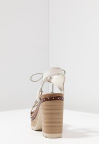 Högklackad sandal med en ljusbeige tygöverdel, brun plattform och strukturerad träklack, prydd med silvernitar längs kanten.