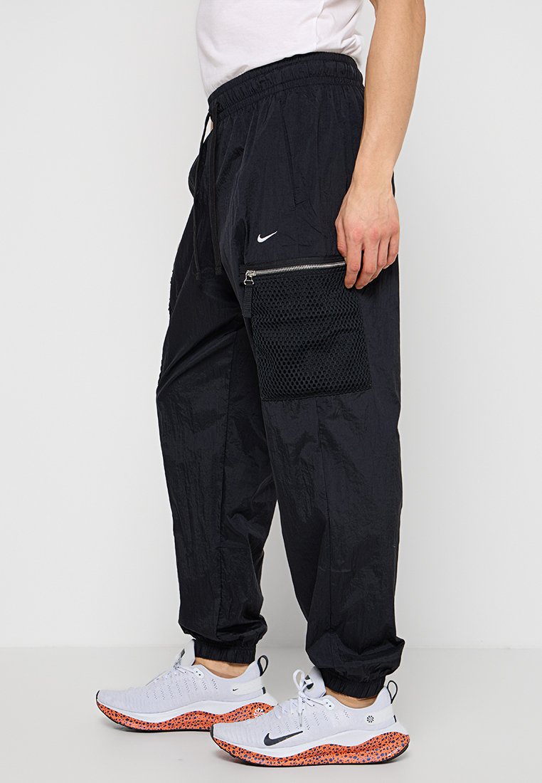 Pantaloni jogger leggeri neri con cintura elastica, tasca laterale con zip dotata di dettagli in rete, abbinati a sneakers bianche con accenti arancioni.