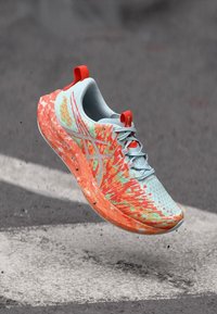 Αθλητικό παπούτσι με επάνω μέρος από ανοιχτό μπλε δίχτυ, ζωντανές πορτοκαλί και πράσινες λεπτομέρειες, και σχέδιο στο πέλμα. Διαθέτει ασφαλές σύστημα σκοινιού.