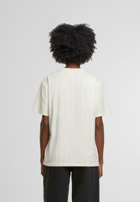 Camiseta de color beige claro con mangas cortas, que presenta sutiles rayas verticales blancas. La parte posterior tiene un escote redondeado y un corte holgado.