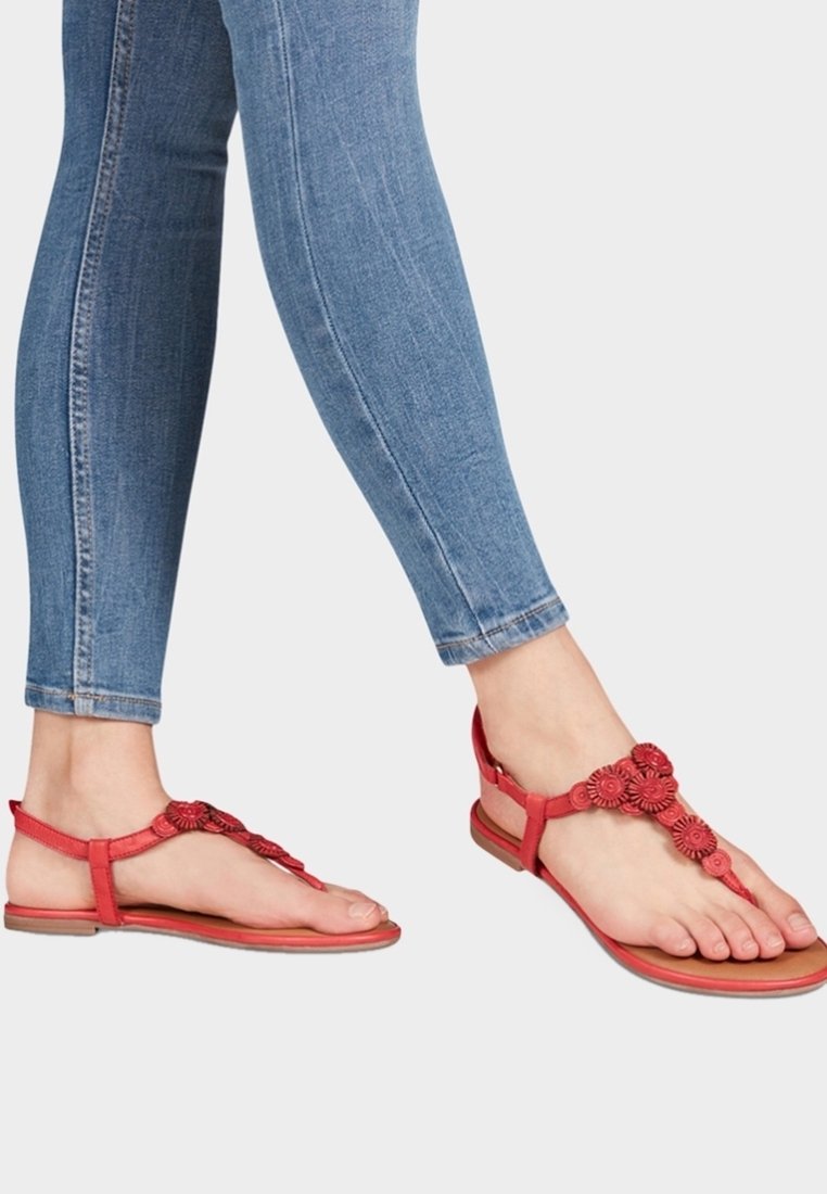Chanclas rojas con acentos de flores; hechas de material sintético. Suela plana, diseño de punta abierta, combinadas con jeans skinny azules.