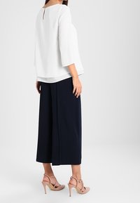 Weiße Bluse mit Viertellangen Ärmeln, Rückenschlitzdetail; kombiniert mit navyblauen weit geschnittenen Hosen und beigen High-Heels.