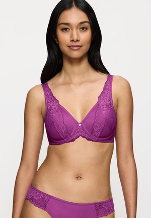 Woman with long black hair wearing matching purple lace bra and panties, standing against a plain light background.