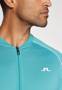 Turkizna športna zip-up majica z visokim ovratnikom, opremljena z črnim zadrgo in belimi akcentnimi liniami. V prednjem delu je majhna logotip.