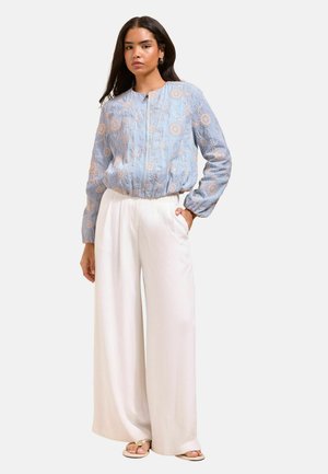 Femme portant une veste zippée fleurie bleu clair et un pantalon blanc large avec des sandales beiges, debout devant un fond uni.