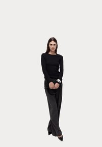 Schwarzes langärmliges Oberteil kombiniert mit grauen weit geschnittenen Hosen. Das Model hält eine schwarze strukturierte Handtasche, trägt spitze schwarze Schuhe und ein silbernes Armband.