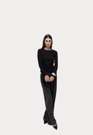 Zwarte lange mouwtop in combinatie met grijze wijde broek. Model houdt een zwarte gevarieerde handtas vast, met puntige zwarte schoenen en een zilveren armband.