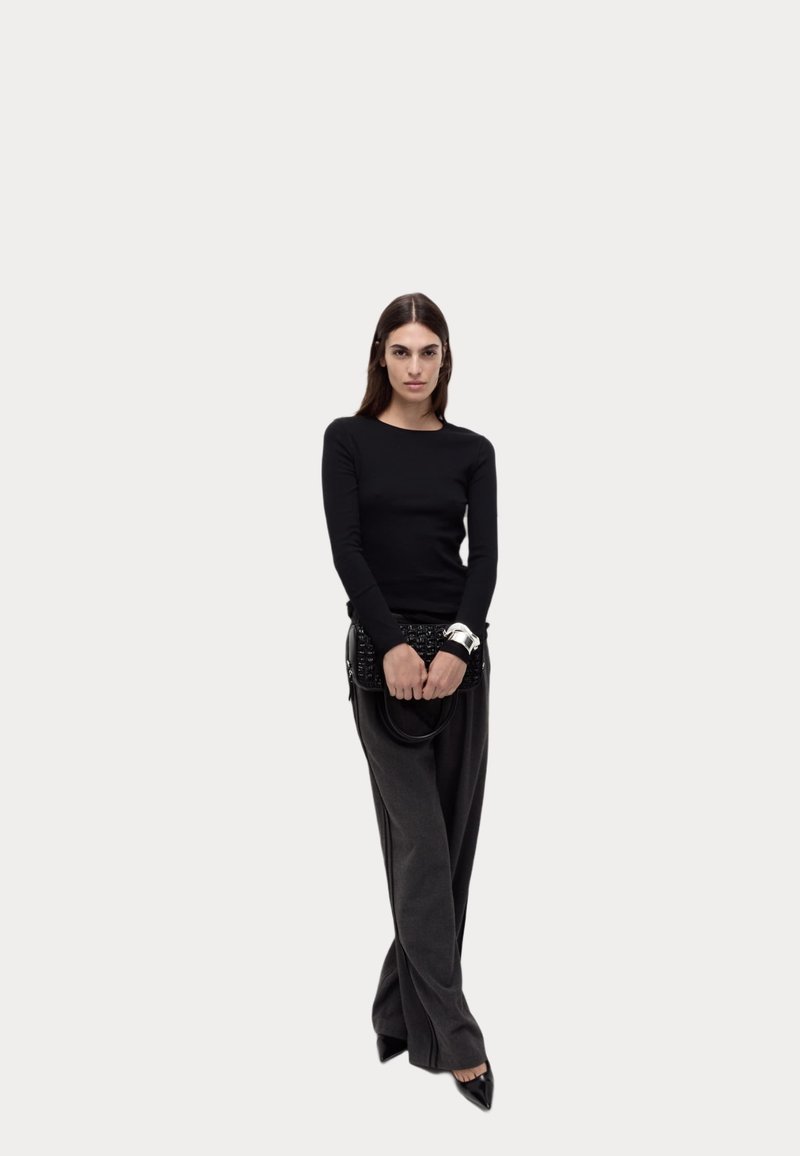 Schwarzes langärmliges Oberteil kombiniert mit grauen weit geschnittenen Hosen. Das Model hält eine schwarze strukturierte Handtasche, trägt spitze schwarze Schuhe und ein silbernes Armband.