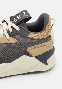Zapatilla deportiva con parte superior en gris y marrón claro, detalles en malla, suela color crema y texto impreso "RS Running System" en el lateral.