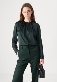 Mörkgrön blus med långa ärmar och veckad halsringning, matchad med byxor i samma nyans och en liten svart handväska. Slät, glänsande tygstruktur.