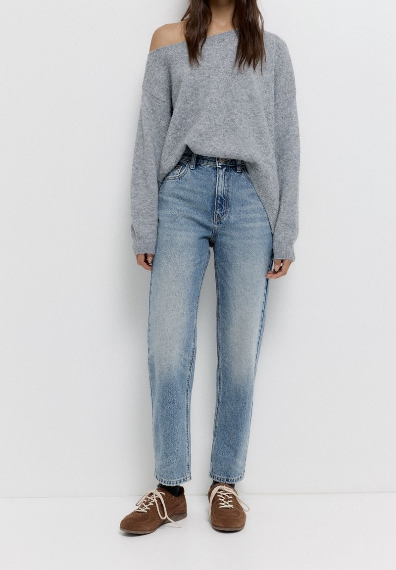 Personne portant un pull gris ample décolleté aux épaules, un jean bleu clair taille haute et des chaussures marron à lacets, debout contre un mur blanc.