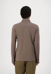 Camisa de manga longa castanha com colarinho, apresentando uma textura suave e punhos com botões. A vista traseira mostra um design limpo com costuras nos ombros.