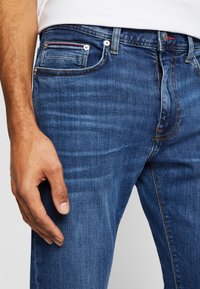 Hombre vestido con jeans de mezclilla azul y una camisa blanca, con su mano izquierda descansando junto a su muslo contra un fondo liso.
