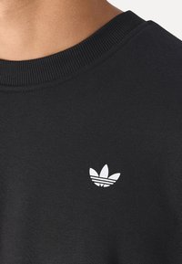 Schwarzer Sweatshirt mit einem gerippten Kragen und einem kleinen weißen Logo mit drei Blättern auf der Brust. Der Stoff wirkt weich und strukturiert.